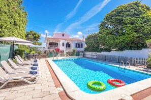 A swimming pool with lounge chairs, a wooden fence, and a building in the background.