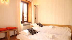 A bedroom with a wooden bed, a bench, a window with blinds, and a floor lamp. - Fantastic location in the heart of Chamonix, a large studio great option for couples or families (Chamonix-Mont-Blanc)