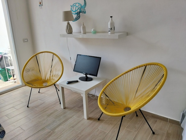 Une pièce avec deux fauteuils en osier jaune, un bureau blanc avec un écran d’ordinateur, et une étagère au-dessus contenant une lampe, une tortue décorative et une bouteille.