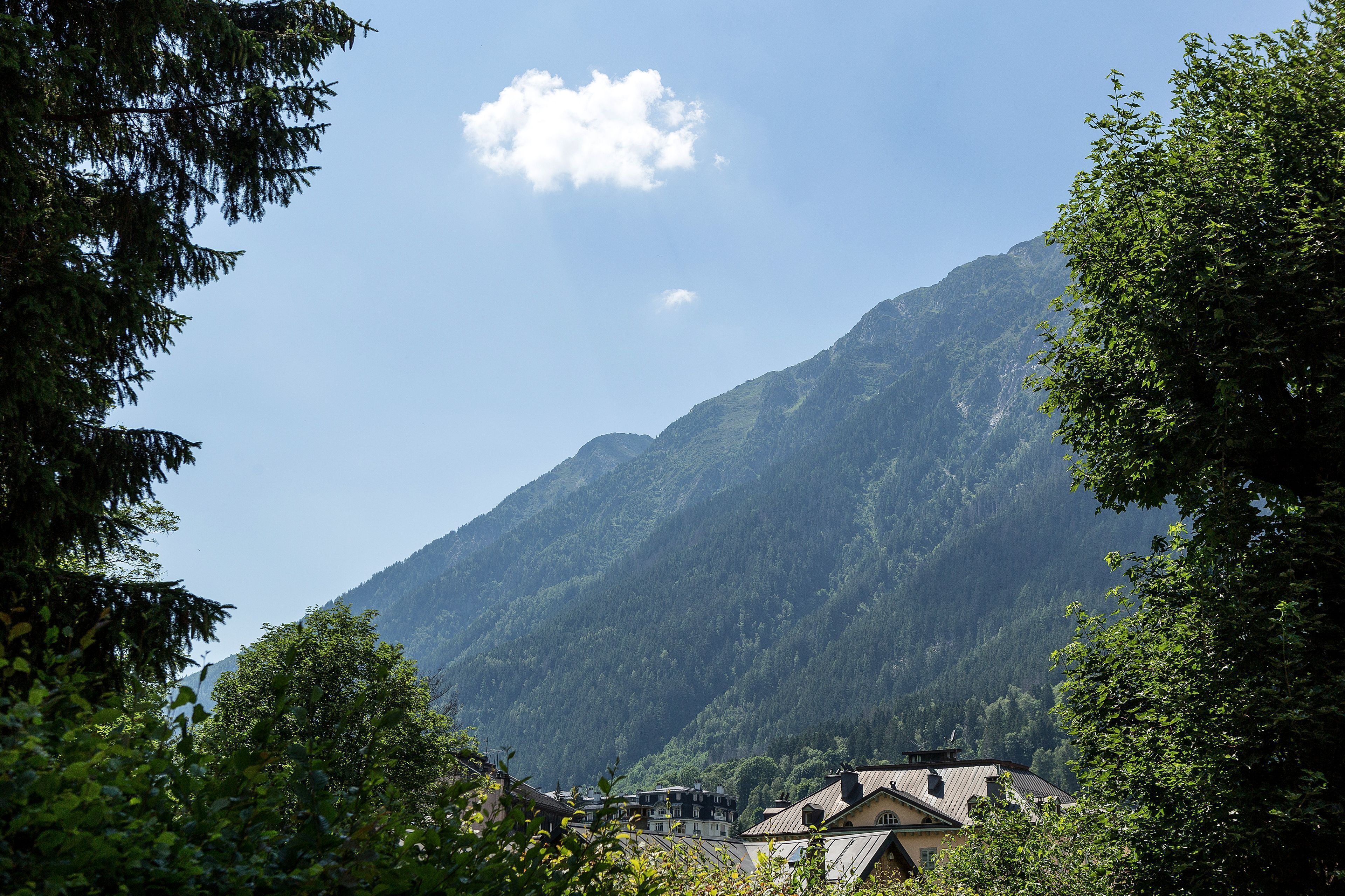 Eine Berglandschaft mit blauem Himmel und vereinzelten Wolken. Am Berghang befinden sich Gebäude, umgeben von Bäumen.