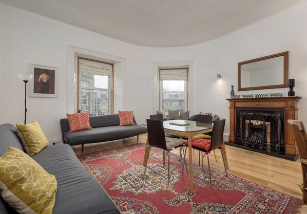 Una sala de estar con un sofá gris, un tapete rojo estampado, una chimenea y una mesa de comedor con sillas.