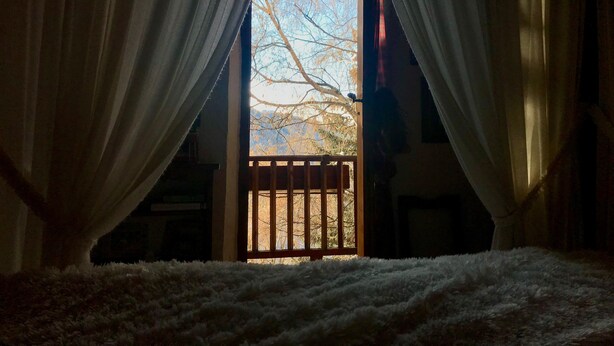 Una habitación con un ventanal grande cubierto por cortinas, una barandilla de madera en el exterior y una alfombra blanca y mullida en el suelo.