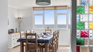 A dining area with a view of the sea, wooden chairs, a table with a tablecloth, and a decorative lamp. - 3 Bed in Woolacombe (oc-1devo) (Woolacombe)