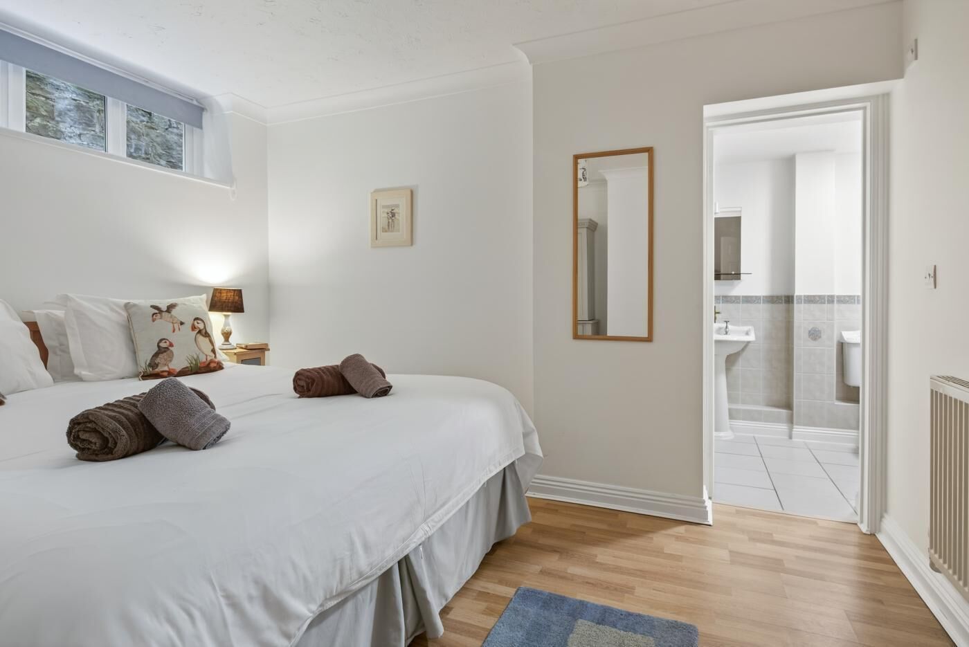 A bedroom with a bed, pillows, a bedside table, a mirror, and a bathroom visible through an open door.