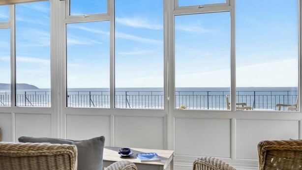 A room with large windows overlooking a beach, wicker furniture, and a table with a cup and saucer. - 3 Bed in Woolacombe (oc-1devo) (Woolacombe)