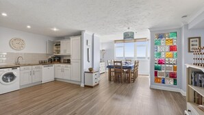 A modern kitchen with white cabinets, a built-in washing machine, and a dining area with a view. - 3 Bed in Woolacombe (oc-1devo) (Woolacombe)