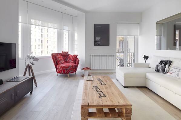 A modern living room with a large window, a red patterned armchair, a white sofa, a wooden coffee table, and a television on a stand. - Zurriola Zinema by FeelFree Rentals (San Sebasti��n)