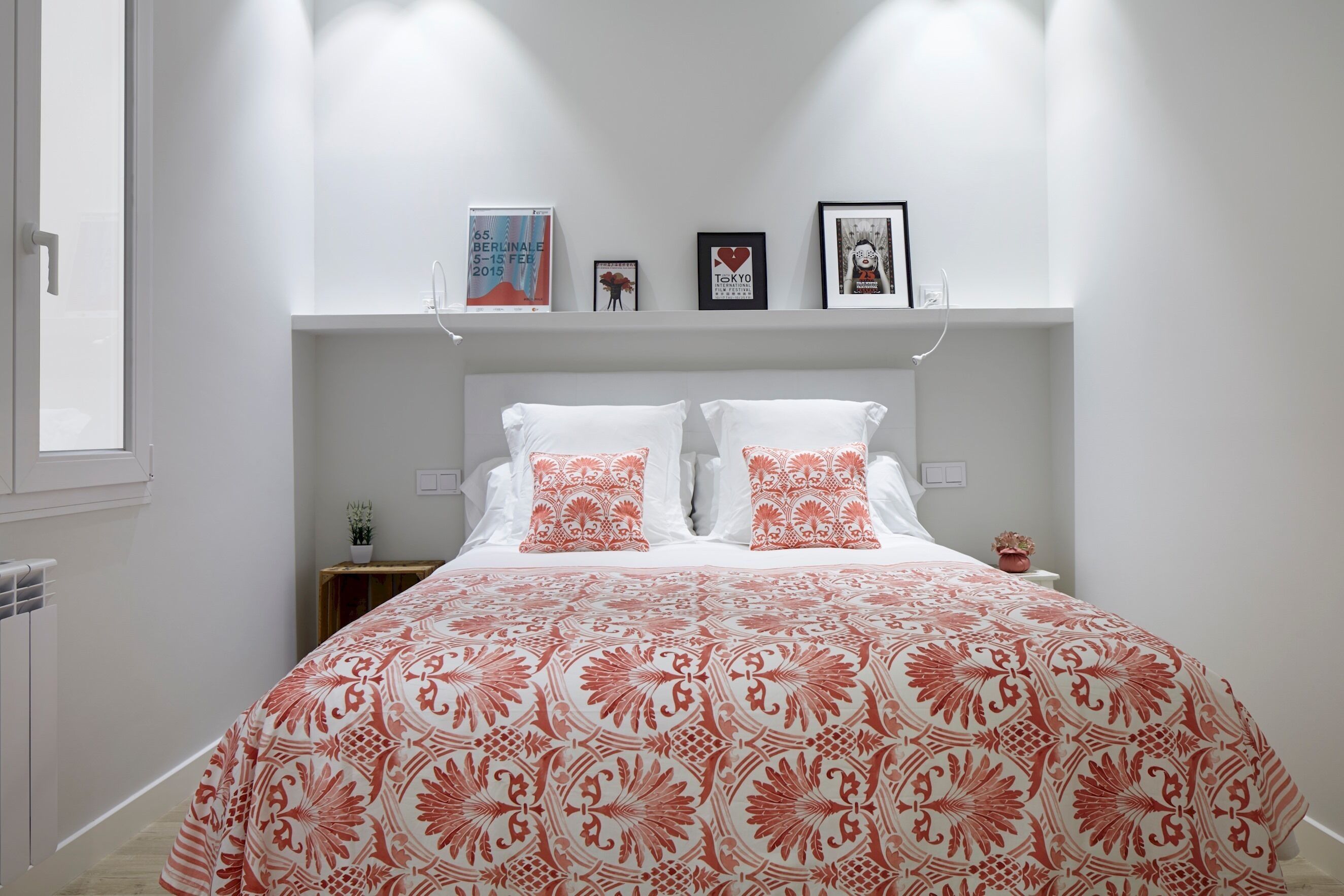 Une chambre avec un grand lit, des murs blancs et une étagère où sont exposés des tableaux encadrés.