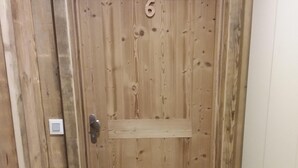 Uma porta de madeira com o número '6' acima, um banquinho pequeno e um tapete com a palavra 'Bienvenue', e um interruptor de luz na parede.