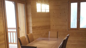 Uma sala de jantar ampla com uma mesa de madeira grande e seis cadeiras, um ventilador de teto e janelas grandes que permitem entrada de luz natural.