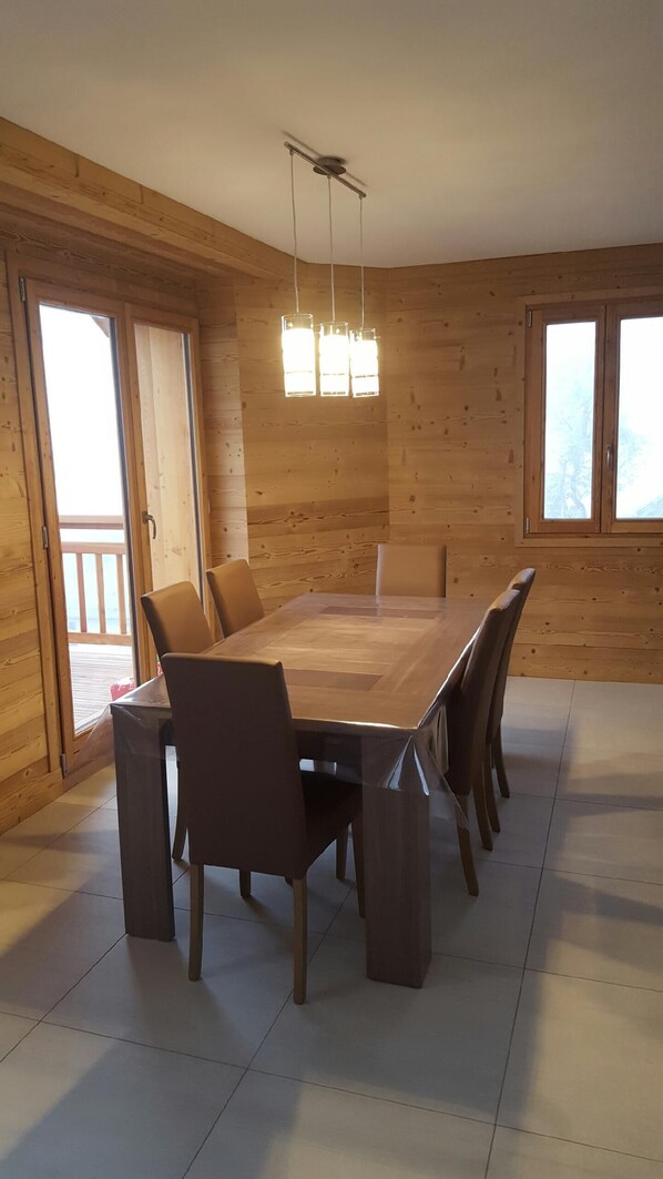 Un comedor amplio con una gran mesa de madera y seis sillas, un ventilador de techo y ventanas grandes que dejan entrar luz natural.