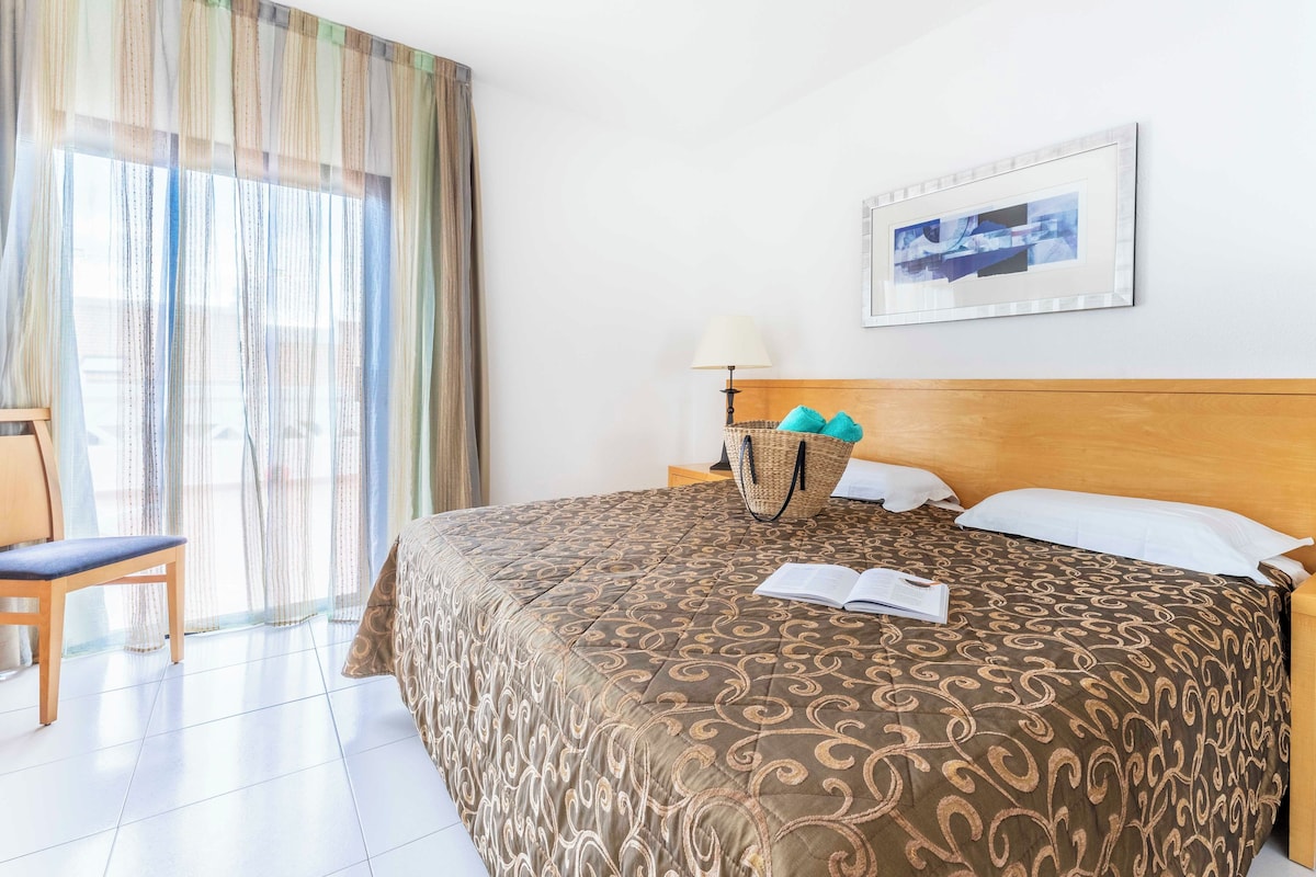 A hotel room with a bed, a chair, a bedside table with a lamp, a window with curtains, and a framed picture on the wall.