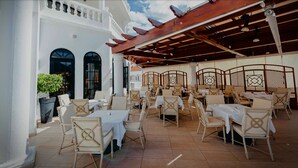 Un café al aire libre con mesas y sillas blancas, una pérgola de madera y un edificio blanco con ventanas arqueadas.