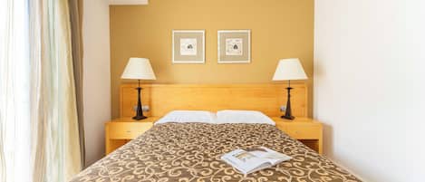 A hotel room with a bed, two bedside lamps, a nightstand, and two framed pictures on the wall.