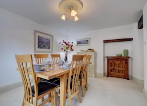 A dining room with a wooden table set for six, surrounded by six wooden chairs with black cushions. There is a white cabinet with a vase of flowers on top, and a wooden cabinet with decorative items on the right.