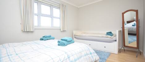 A bedroom with a bed, a window with curtains, a bedside table, a mirror, and a wooden floor.