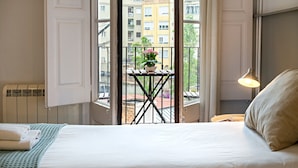 Habitación de hotel con una cama, una ventana que ofrece vistas a edificios, una mesita con una planta y una lámpara.