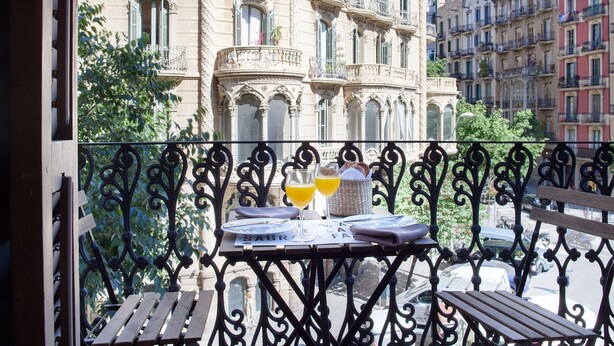 Un balcón con una mesa dispuesta para dos personas, que incluye una cesta, dos vasos de zumo de naranja y una botella.