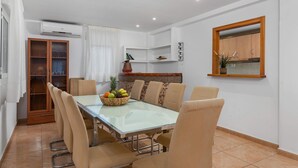 A dining area with a glass table surrounded by chairs, a fruit bowl on the table, and a kitchen area with cabinets and a window. - Villa Caleta - PlusHolidays (Calpe)