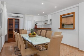 A dining area with a glass table surrounded by chairs, a fruit bowl on the table, and a kitchen area with cabinets and a window. - Villa Caleta - PlusHolidays (Calpe)