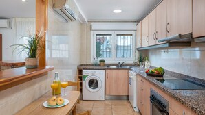 A modern kitchen with a built-in washing machine, a dining area with a table set for two, and a window overlooking a balcony. - Villa Caleta - PlusHolidays (Calpe)