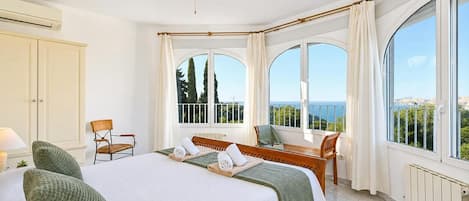 A hotel room with a bed, two chairs, a table, and large windows offering a view of the sea.