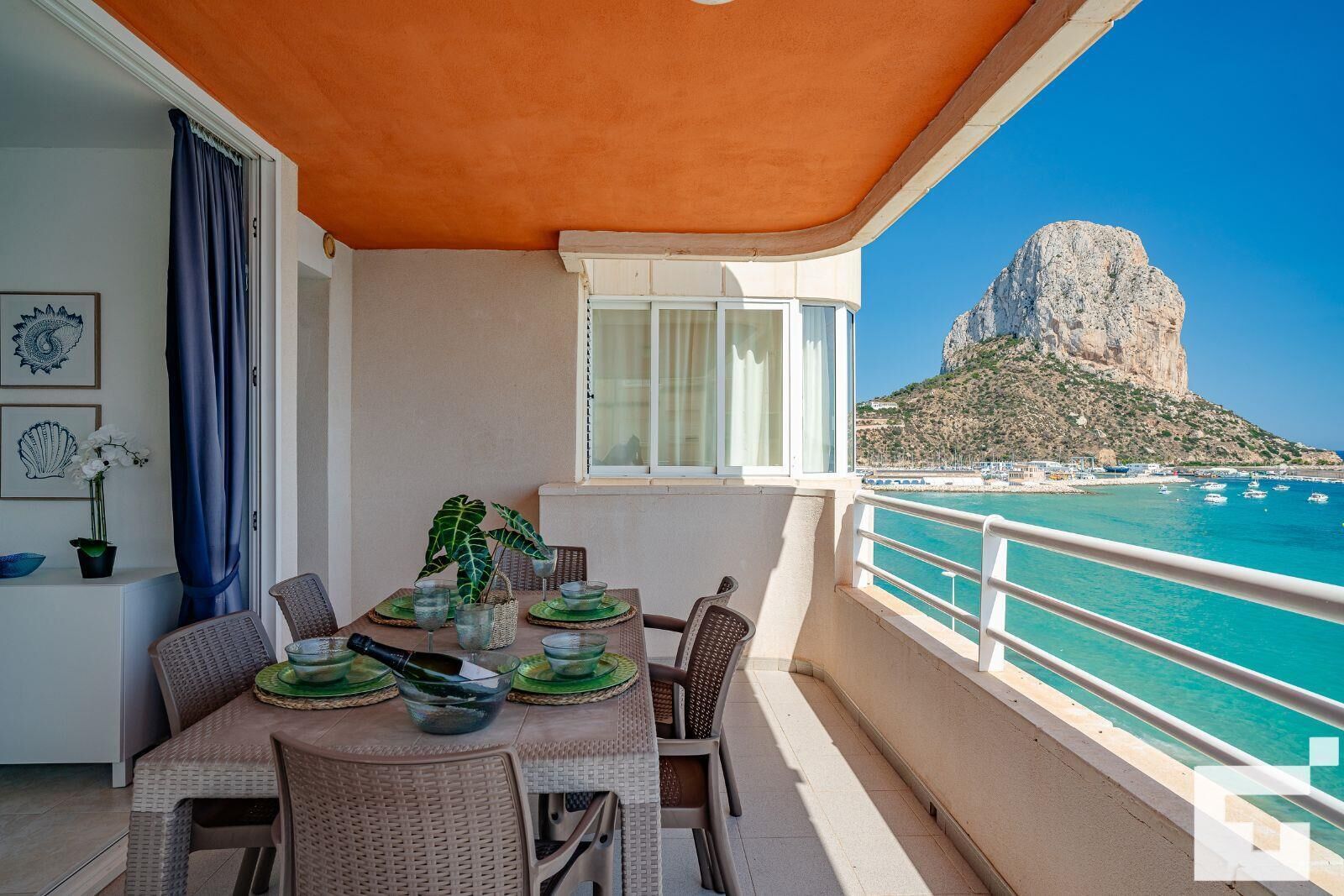 Un balcón con mesa lista para comer, con vistas a la costa y una destacada formación rocosa.