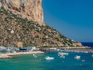 A coastal marina with boats docked, a rocky cliff, and buildings along the waterfront.