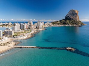 An aerial view of a coastal city with a prominent rocky hill, clear blue waters, and a mix of residential and commercial buildings along the shore.