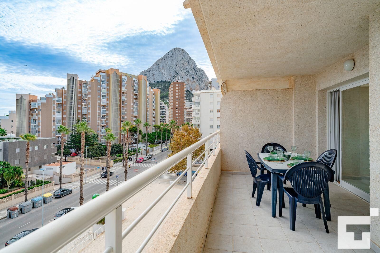 Un balcón con una mesa y sillas, con vista a una calle con coches y edificios, y una montaña al fondo.