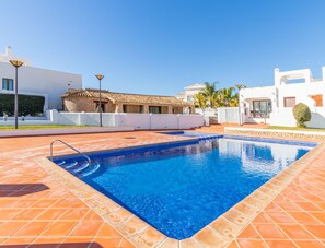 Una piscina con borde de baldosas, rodeada de edificios blancos y un muro de piedra.
