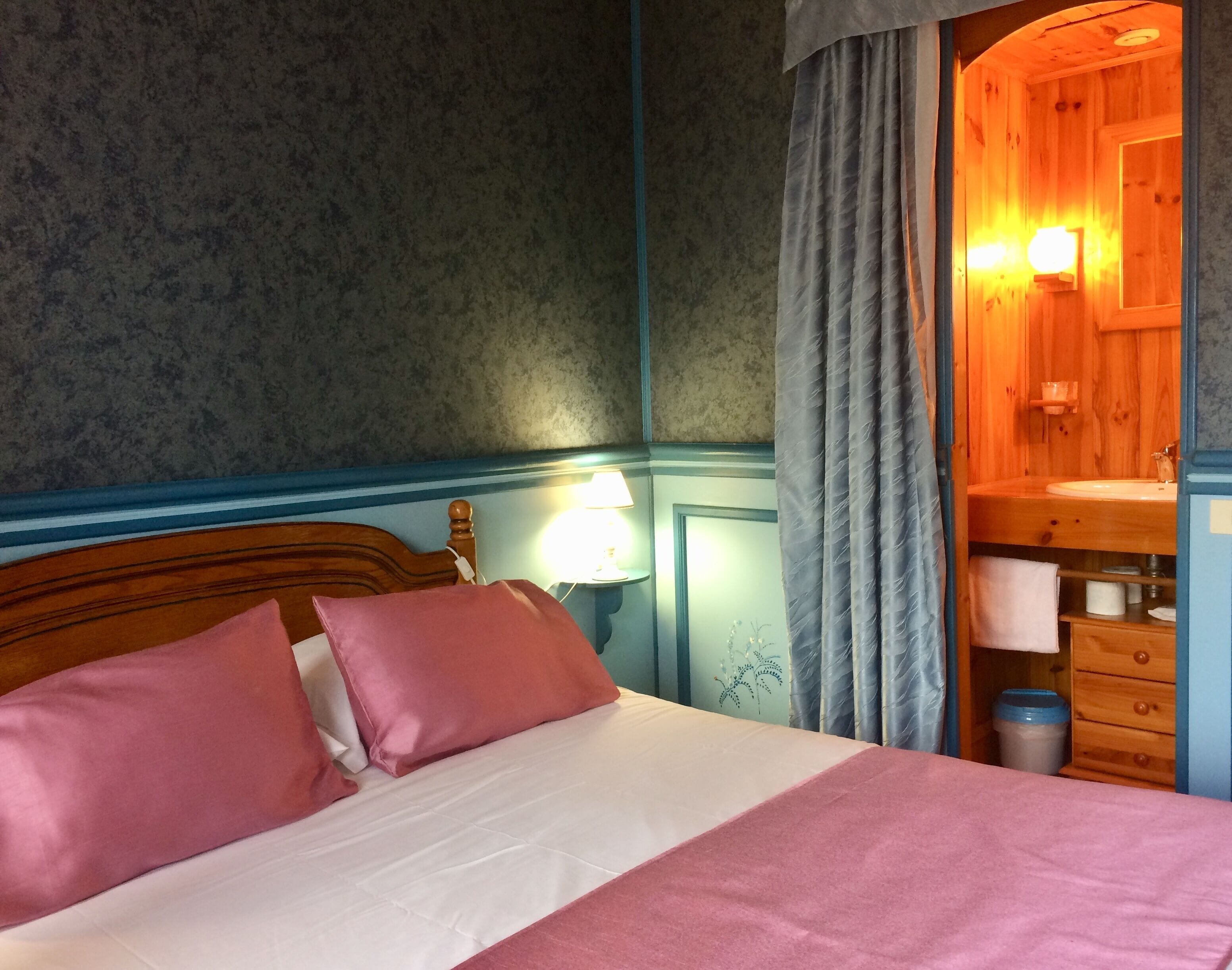 Un dormitorio con cabecera de madera, una cama con ropa de cama rosa y blanca, una lámpara de noche y un mueble de madera con lavamanos.