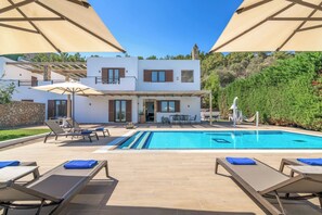 A pool area with lounge chairs and umbrellas, a pool, and a building with a stone facade and wooden shutters.