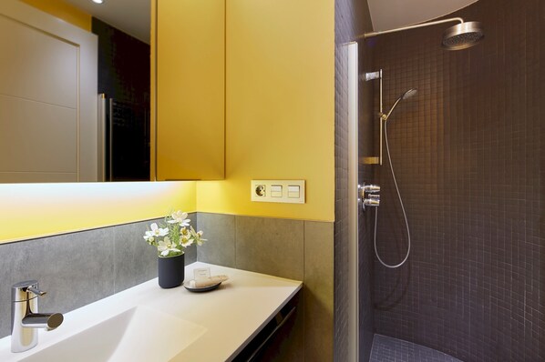 Une salle de bain moderne avec un lavabo blanc, une plante en pot et un espace douche avec un mur recouvert de carreaux foncés.