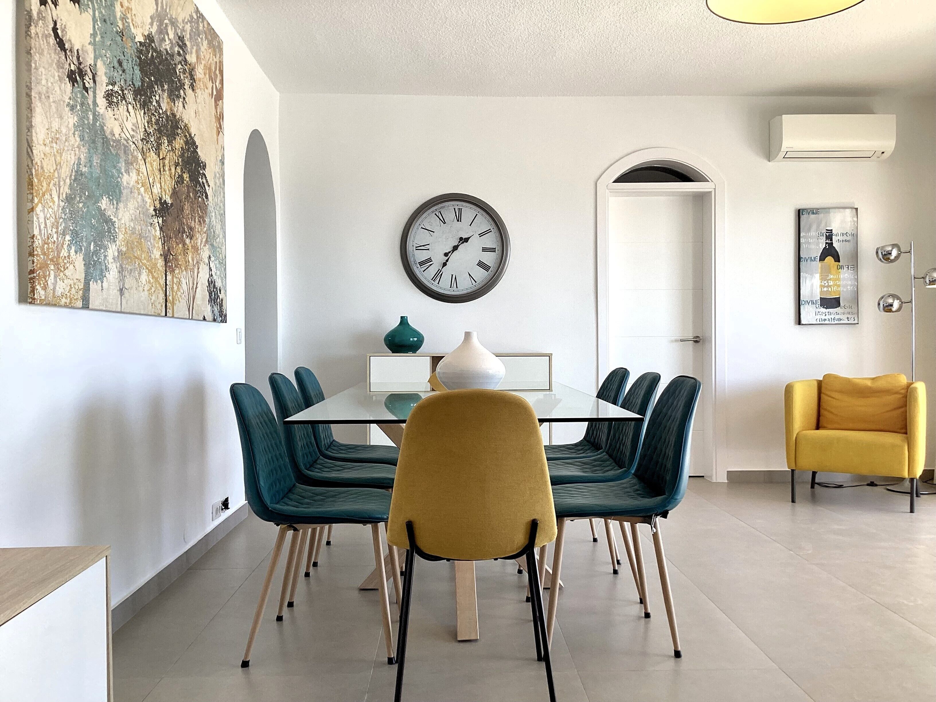 Een eethoek met een glazen tafel, teal bekleden stoelen en een gele stoel. Op de muur hangt een klok, en boven de tafel een schilderij.