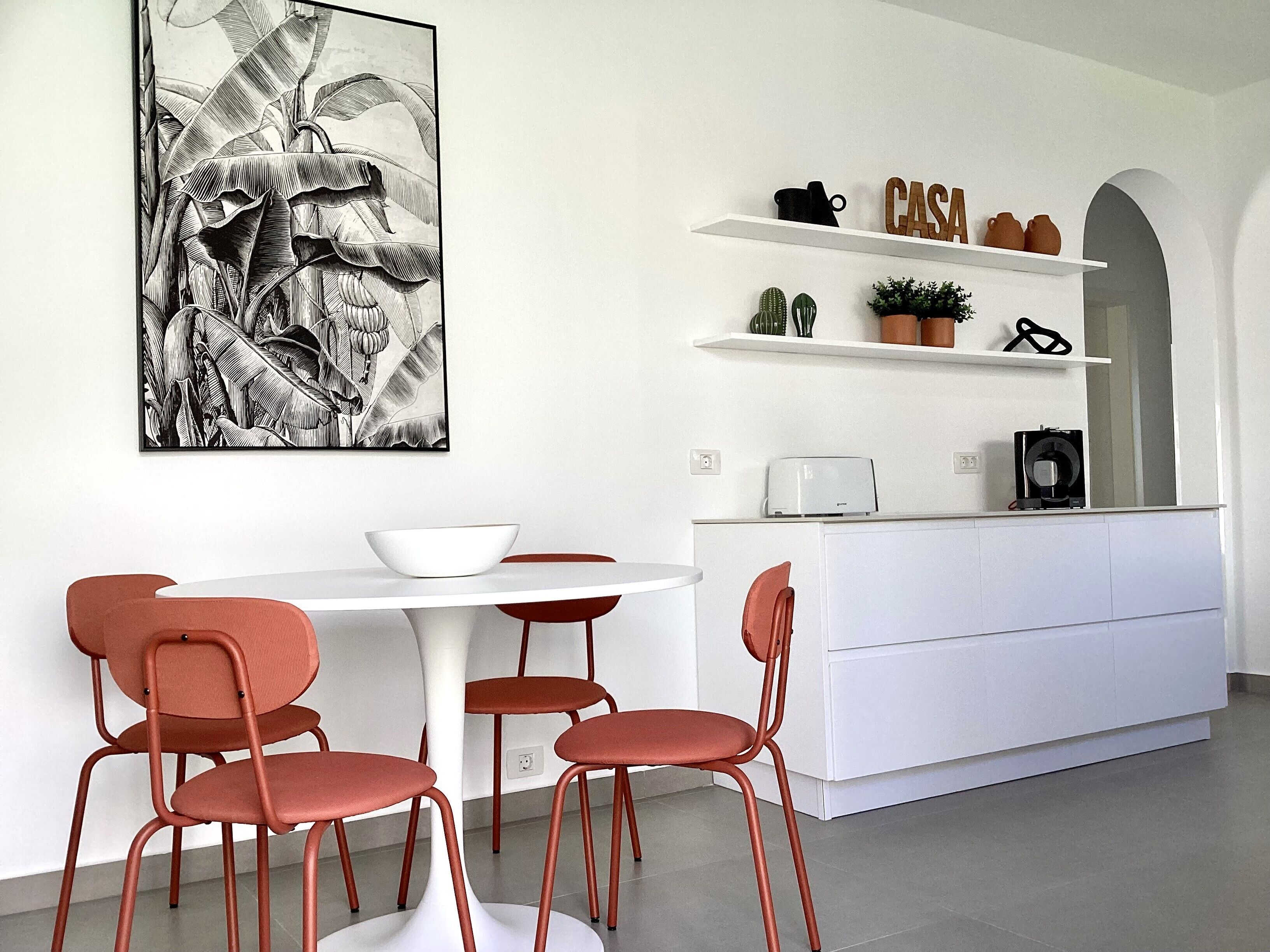 Una cocina moderna con una mesa de comedor blanca, cuatro sillas rojas, un armario blanco y una obra de arte blanco y negro en la pared.