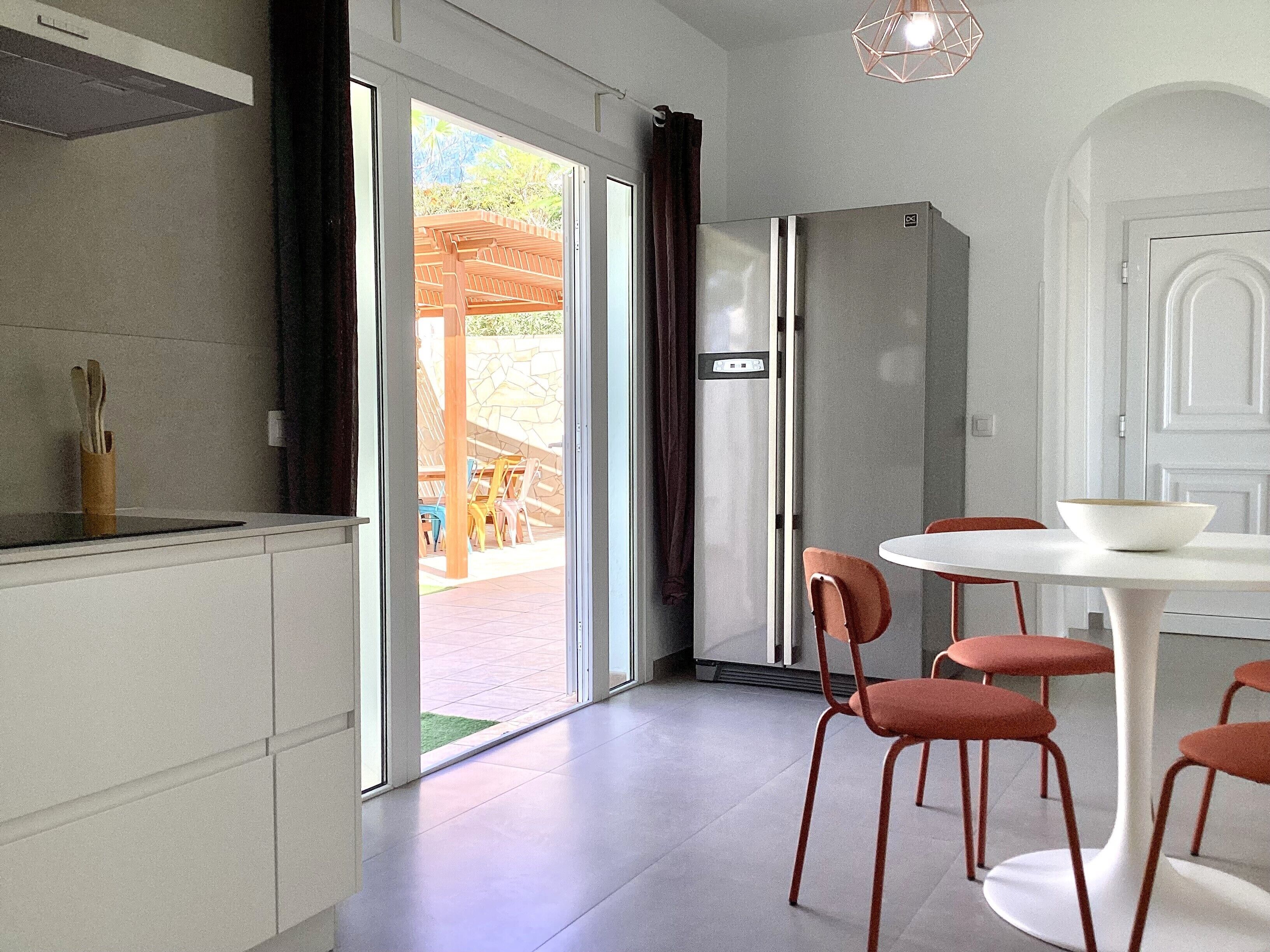 Eine moderne Küche mit Backofen, ein Essbereich mit weißem Tisch und roten Stühlen sowie eine Glastür, die auf eine Terrasse im Freien führt.