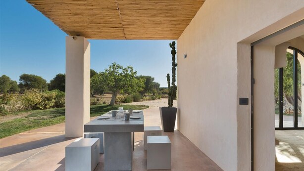 Une terrasse extérieure avec une table dressée pour le repas, entourée de piliers blancs, d’un cactus et offrant une vue sur le paysage.