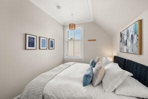 A bedroom with a bed, framed artwork, a window with blinds, and a ceiling light.