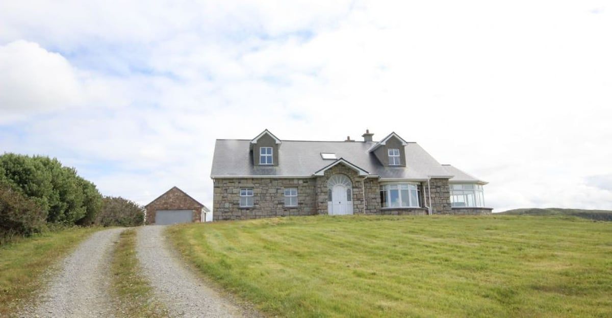 A stone house with a gravel driveway leading to a garage, surrounded by grass and trees.