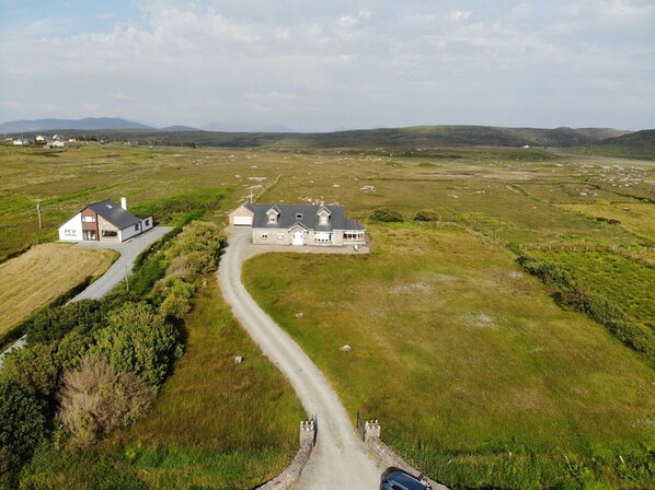 A large, two-story house with a driveway leading to a gated entrance, surrounded by open fields and distant hills. - Jackie's Cottage, overlooks Omey Island & strand (Claddaghduff)