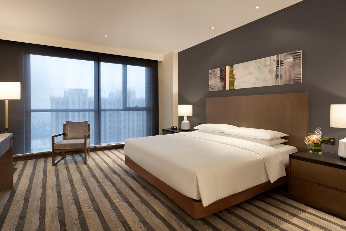 Habitación de hotel con una cama grande, una silla, un escritorio con televisor y una ventana con vista a la ciudad.