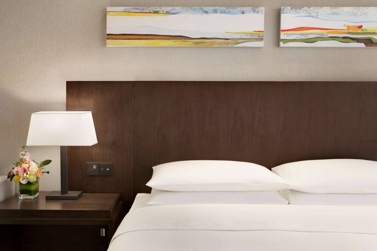 Een hotelkamer met een houten hoofdbord, een nachtkastje met een lamp en bloemen, en twee schilderijen aan de muur.