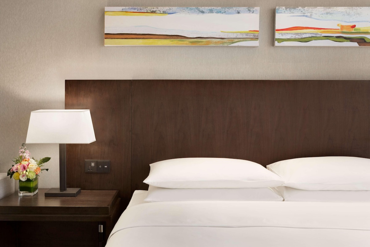 Una habitación de hotel con una cama, una mesita de noche con una lámpara y flores, y dos cuadros abstractos en la pared.