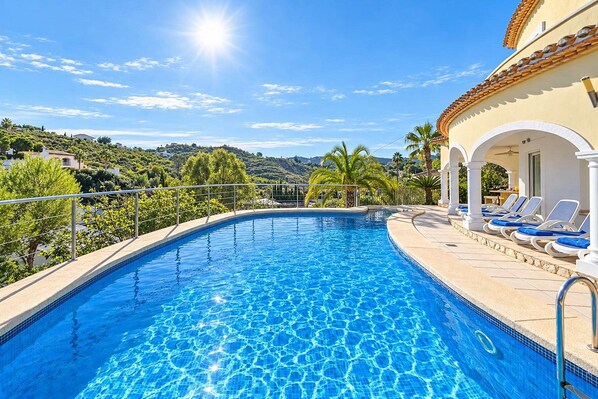 A swimming pool with clear blue water, surrounded by lounge chairs and a building with a tiled roof. - Villa Jarmila - Javea, Costa Blanca (Javea)
