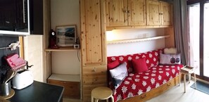 Una habitación acogedora con un armario de madera, una cama con colcha floral roja, un taburete pequeño y una ventana con cortinas.