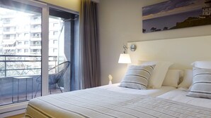 Quarto de hotel com uma cama, mesa de cabeceira, luminária, cadeira e vista para um edifício pela janela.