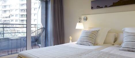 A hotel room with a bed, a nightstand, a lamp, a chair, and a view of a building through a window.