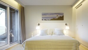 Quarto com uma cama grande, duas luminárias de cabeceira, um quadro de um farol, uma porta de correr de vidro e uma cadeira perto da janela.