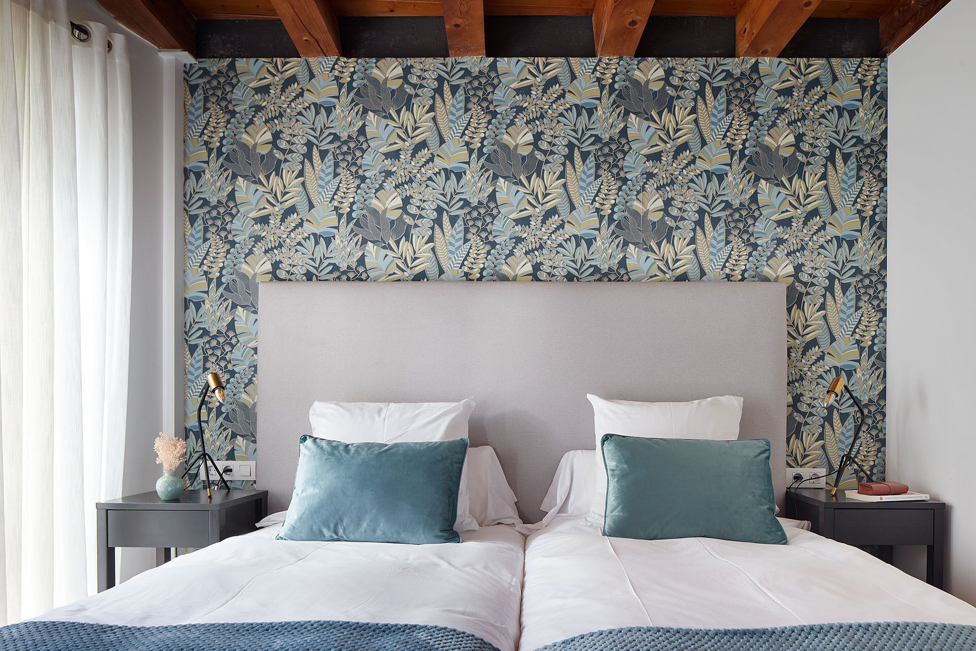 Un lit double avec une literie blanche et turquoise, une tête de lit grise et un papier peint à motifs floraux.